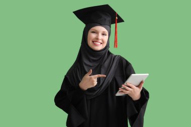Yeşil arkaplanda tablet bilgisayarına işaret eden Müslüman mezun öğrenci