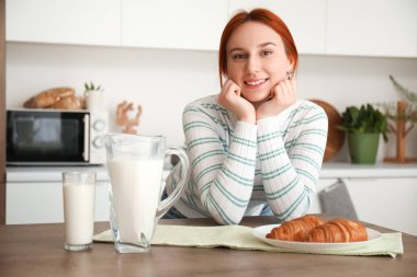 Mutfakta bir bardak süt ve kruvasanla genç kızıl saçlı kadın.