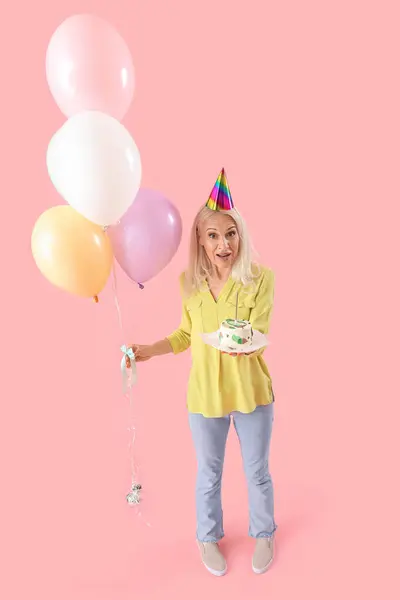 Mature woman with birthday cake and balloons on pink background