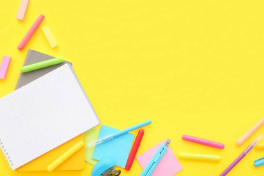 Boş defterli kompozisyon ve sarı arka planda farklı okul malzemeleri.