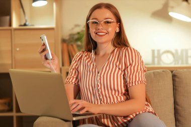 Akşam evde cep telefonu ve dizüstü bilgisayarla çalışan genç bir kadın.