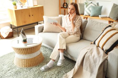 Evde cep telefonuyla konuşan tablet bilgisayarlı çalışan genç bir kadın.