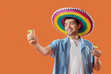 Sombrero şapkalı genç adam, turuncu arka planda tekila shot atıyor.