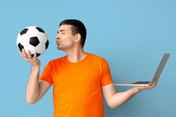 Happy young man with soccer ball and laptop on blue background. Sports bet concept