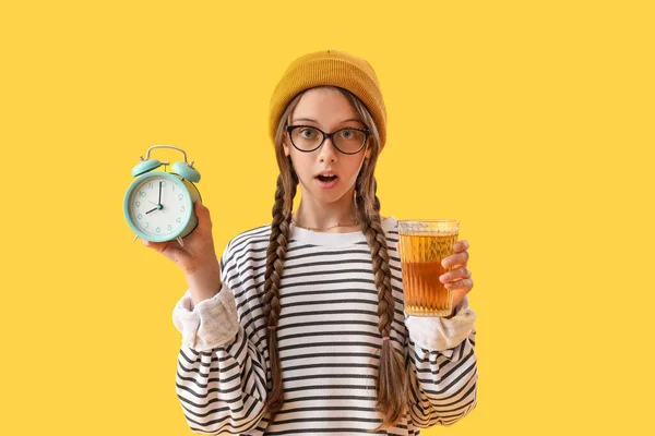Shocked little girl with glass of juice and alarm clock on yellow background