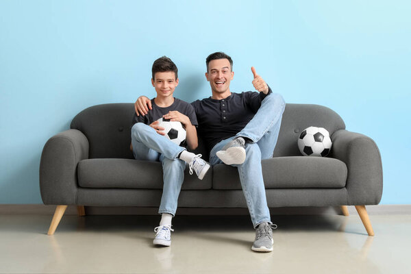 Little boy with his dad watching football game on sofa near blue wall