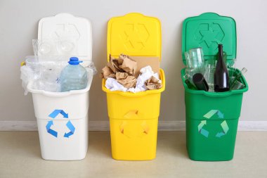 Geri dönüşüm sembollü çöp kutuları ve beyaz duvarın yanındaki farklı çöpler.