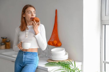 Bir fincan kahveyle genç ve güzel bir kadın mutfakta lezzetli kruvasan yiyor.