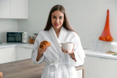 Lezzetli kruvasan ve mutfakta bir fincan kahveyle bornozlu güzel bir kadın.