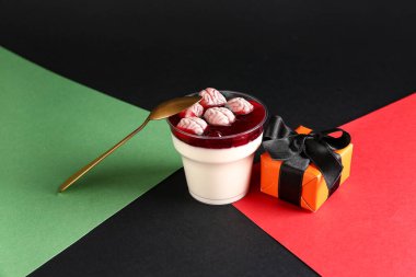 Renkli arkaplanda Cadılar Bayramı kutlaması için lezzetli panna cotta ve hediye kutusu olan plastik bardak, yakın plan.