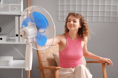Oturma odasında elektrikli yelpaze ile oturan güzel, mutlu genç bir kadın.