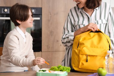 Anne, küçük oğluna okul çantasını ve öğle yemeğini hazırlaması için mutfakta yardım ediyor.