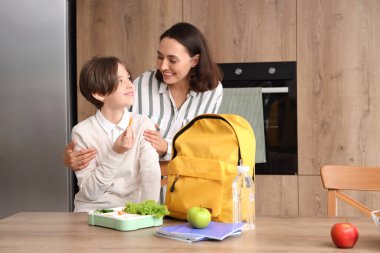 Anne, küçük oğluna okul çantasını ve öğle yemeğini hazırlaması için mutfakta yardım ediyor.