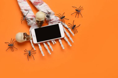 Modern cep telefonlu iskelet eller, turuncu arka planda Cadılar Bayramı kutlaması için örümcekler ve kafatasları.