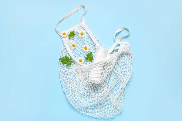 Mesh bag with beautiful chamomile flowers and bottle of cosmetic product on blue background