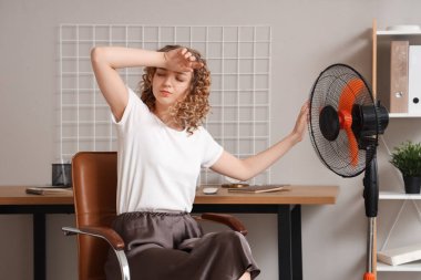 Evdeki sıcak hava dalgası sırasında elektrikli yelpazeli güzel bir kadın sandalyede oturuyor.
