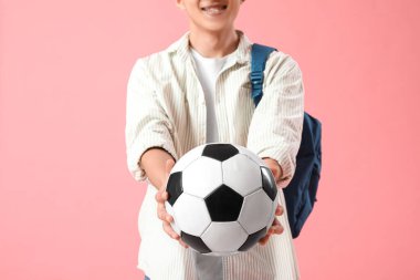 Sırt çantası ve pembe arka planda futbol topu olan mutlu Asyalı erkek öğrenci.