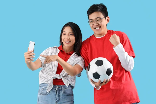 Young Asian friends with mobile phone and soccer ball on blue background