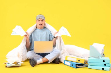 Stresli, pijamalı, evrakları ve laptopu olan genç adam sarı arka plan üzerinde çalışıyor.