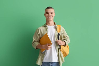 Yeşil arka planda not defterleri ve sırt çantası olan mutlu bir erkek öğrenci.