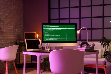 Karanlık ofiste bilgisayar, dizüstü bilgisayar ve parlayan lambayla programcının çalışma yeri