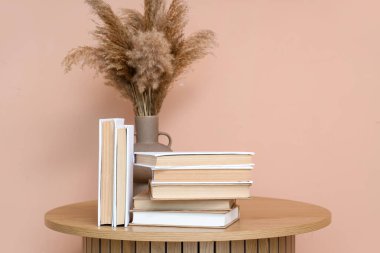 Bej duvarın yanındaki masada otlu vazo ve kitaplar.