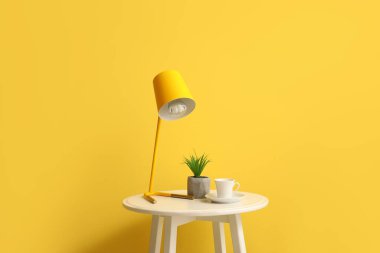 Sarı duvarın yanında lamba, fincan ve çiçek olan küçük bir masa.