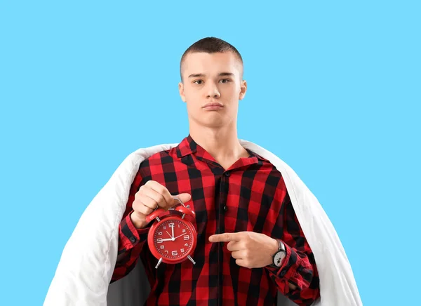 Young man in pajamas, with blanket and alarm clock on blue background
