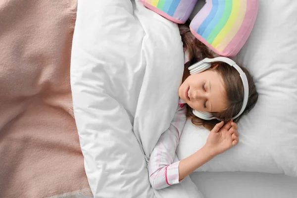 Little girl with headphones sleeping in bed, top view