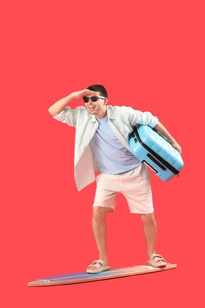 Männlicher Tourist Mit Koffer Steht Auf Surfbrett Vor Rotem Hintergrund — Stockfoto