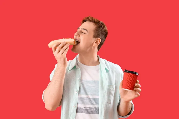 stock image Young man with tasty hot dog and cup of coffee on red background