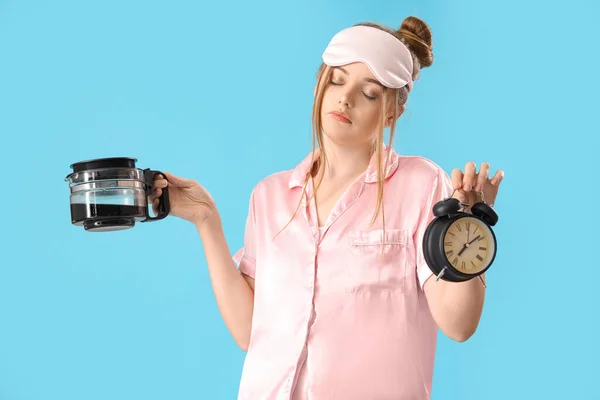 Sleepy young woman with alarm clock and coffee pot on blue background