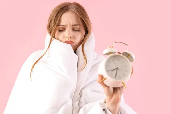 Sleepy young woman with alarm clock and blanket on pink background