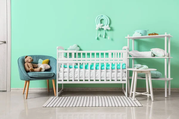 Çocuk odasının içinde, turkuaz duvarın yanında beşik ve koltuk var.