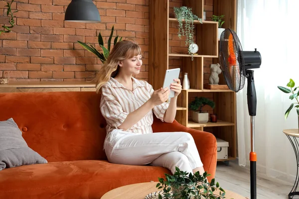 Tablet bilgisayarı ve elektrikli vantilatörü olan genç bir kadın evdeki koltukta oturuyor.