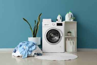 Çamaşır makinesi, raf ünitesi, ev bitkisi ve mavi duvarın yanındaki kirli çamaşır yığını.