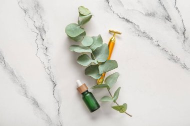 Beyaz zemin üzerinde serum ve okaliptüs yapraklı metal yüz masajı silindiri.