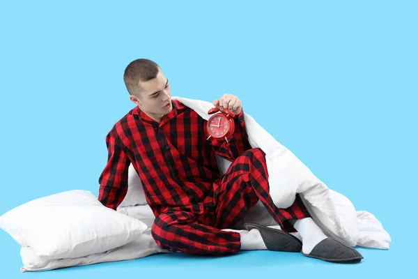 Young man in pajamas and with alarm clock on blue background