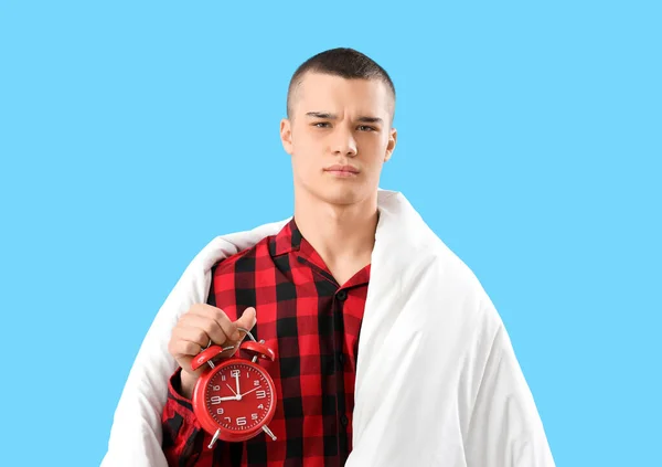 Displeased young man in pajamas, with blanket and alarm clock on blue background