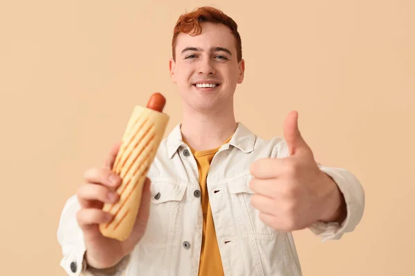年轻男子与美味的热狗显示大拇指向上的姿态米色背景 — 图库照片