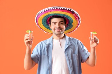 Sombrero şapkalı genç adam, turuncu arka planda tekila shot atıyor.