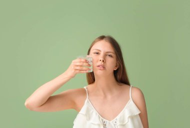 Dişi ağrıyan genç bir kadın elinde yeşil arka planda bir bardak soğuk su tutuyor.