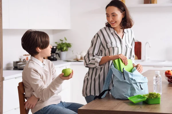 Mother helping her little son to pack schoolbag and lunch during breakfast in kitchen