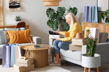 Evde cep telefonu ve yeni alımları olan olgun bir kadın. İnternetten alışveriş