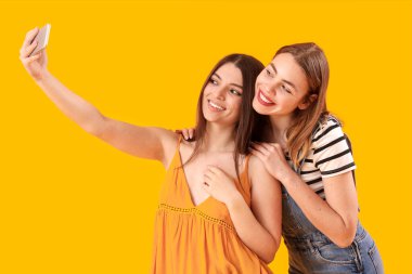 Cep telefonlu bayan arkadaşlar sarı arka planda selfie çekiyorlar.
