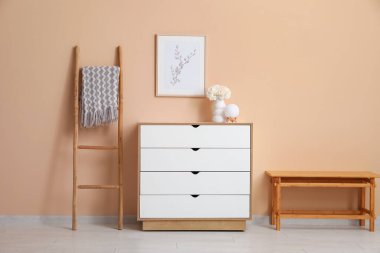 İçinde beyaz şakayık vazosu olan çekmeceler ve bej duvar kenarındaki tablolar.