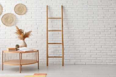 Beyaz tuğla duvarın yanında kurutulmuş pampa vazosu ve merdiveni olan açık ahşap bir sehpa.