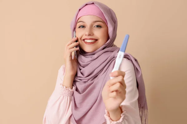 ベージュの背景で電話で話す肯定的な妊娠テストを持つ幸せなムスリムの若い女性 — ストック写真