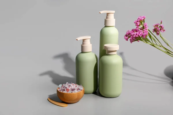 Bottles of cosmetic products with beautiful flowers and sea salt on grey background