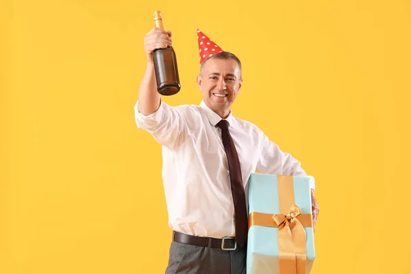 Mature businessman with birthday gift and champagne on orange background